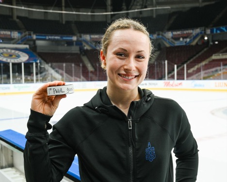 Smiling hockey player stands by the rink holding up a puck toward the camera.
