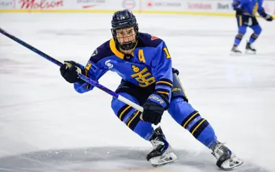 Hockey player in blue uniform skates low to the ice while controlling the puck during a game.