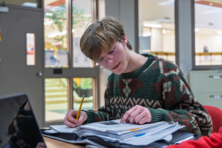 Student working independently at a table, writing in a notebook beside an open binder and laptop.