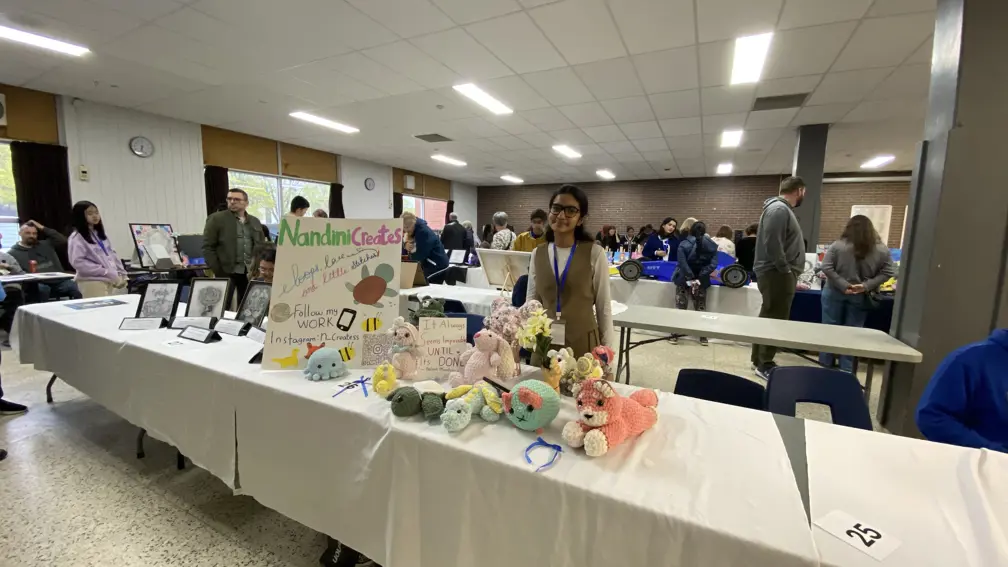 A student stands beside a table displaying handmade crochet crafts and artwork at a school showcase event.