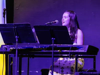 Student sings while playing a keyboard on stage during a live performance under purple lighting