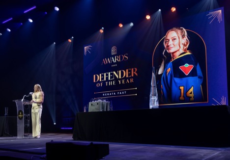 Speaker stands on stage beside a large screen displaying Defender of the Year with a photo of a hockey player in uniform.