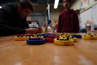 Low-angle view of students working at a table with colorful circular components, one student focused on assembling pieces while another stands nearby in a classroom or workshop setting.