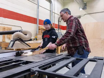 Student wearing safety glasses learning to use a table saw with guidance from a teacher in a woodworking shop.