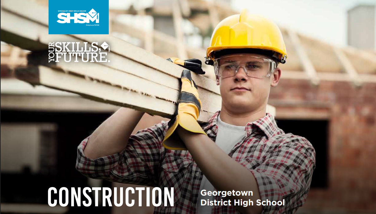 a teenage student wearing a yellow hard hat, safety goggles and work gloves carrying lumber rover their shoulder.