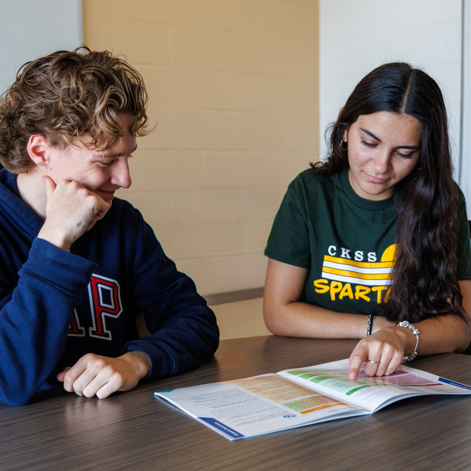 Two students working together with an open textbook at a classroom table.