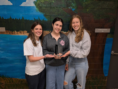 Three students stand together indoors, smiling and holding a trophy in front of a painted mural, celebrating a wetland competition achievement.
