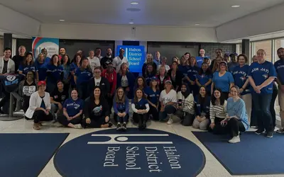 Group of Halton District School Board staff in the lobby wearing Toronto Blue Jays shirts and jerseys, smiling and posing around the HDSB floor logo and sign.