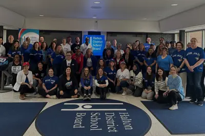 Group of Halton District School Board staff in the lobby wearing Toronto Blue Jays shirts and jerseys, smiling and posing around the HDSB floor logo and sign.