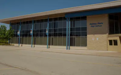 Teacher speaking to high school students in a classroom with large windows and a view of sports fields outside.