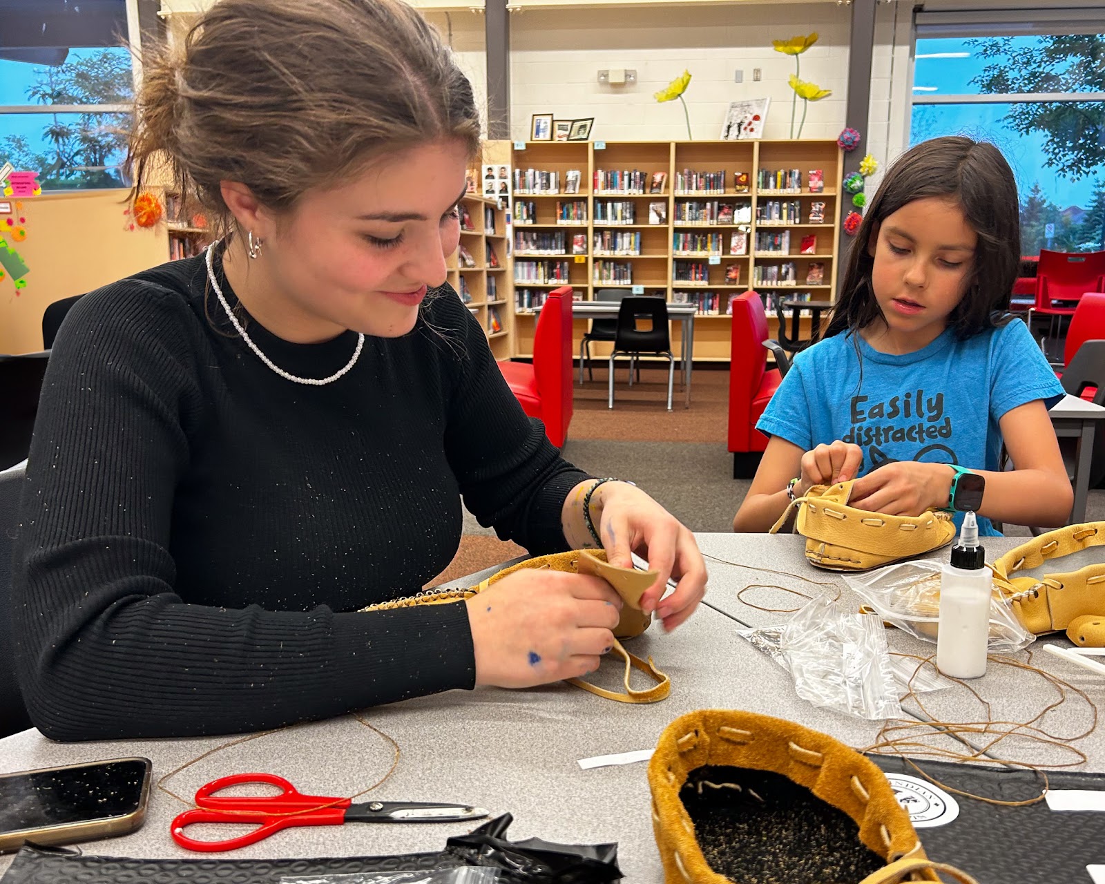 a teenage girl working on a craft in the library with a child.