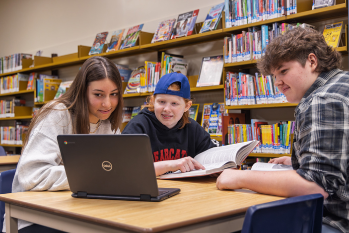 Three students studying together at a library table with a laptop and open books.