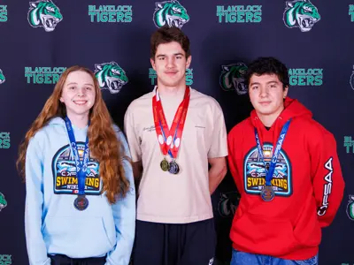 Three students in the photo standing in front of a background that says Blakelock Tigers and has Tiger heads repeated over and over. The girls on the left is wearing a bright blue OFSAA sweatshirt and has a blue ribbon with a medal around her neck. The boy in the middle is wearing a beige tshirt and has three medals, 2 on red ribbons and 1 blue ribbon around his neck. The 3rd boy on the right hand side is wearing a red OFSAA sweatshirt and a medal with a blue ribbon around his neck.