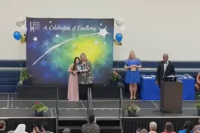 a stage set up at the front of a school gymnasium with one person speaking at a podium and two people standing centre stage. 
