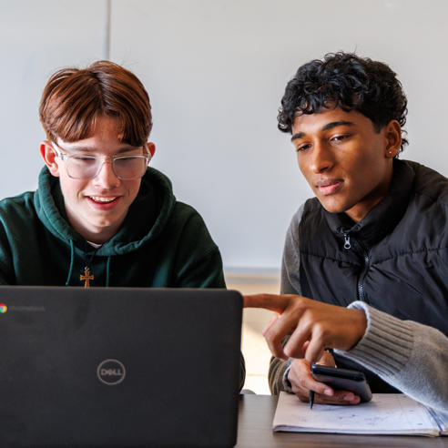 Two students working together on a laptop at a classroom table.