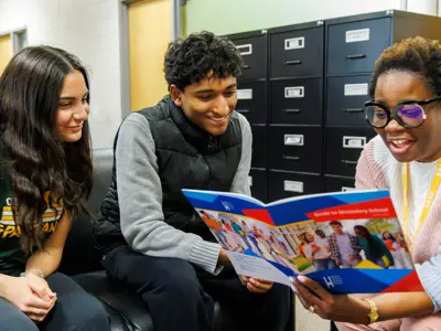 Two students smile as a staff member reads a secondary school guide with them.
