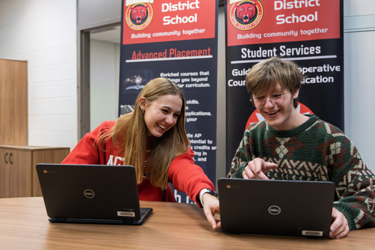 Two students smiling and working together on laptops at a table, with Advanced Placement and Student Services banners behind them