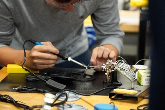 Close-up of a person soldering electronic components on a workbench, with tools, wires, and a helping hands stand visible.