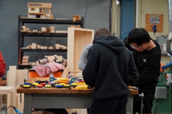 Students working at a table in a woodworking shop, using tools and measuring materials, with shelves of wood and equipment in the background.