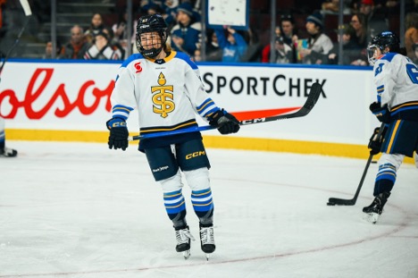 Hockey player in white and blue uniform stands on the ice during gameplay, holding a stick and looking ahead